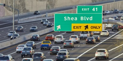 A large road sign is visible from a high angle above a busy highway, directing rush-hour drivers to an exit.

