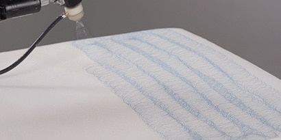 Image of an applicator spraying hot melt adhesive in a consistent side-to-side pattern on a large surface
