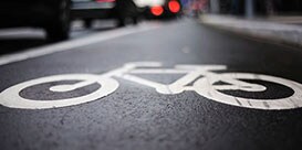 Close-up of biker symbol in white, on black pavement, in urban bike lane with cars in the background.
