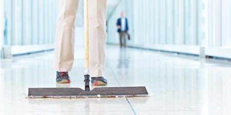 Imagen de una persona de la limpieza limpiando con una mopa plana los residuos del piso del pasillo de una escuela.
