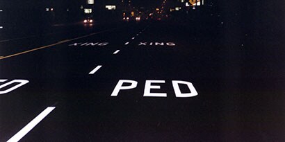 White illuminated pavement markings on dark road at night, reading “PED XING.”

