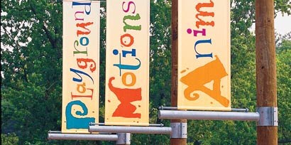 Three wayfinding signs made with 3M grphic films that say playground, motions, and animals on a campground hanging from trees.
