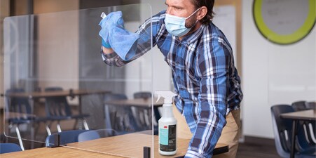 Imagen de un hombre con camisa de tirantes y mascarilla limpia un separador de plástico en un aula.

